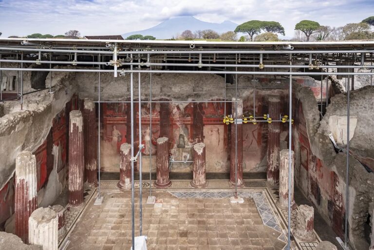 an overview from an elevated perspective of a large-scale fresco in a banquet hall excavated in Pompeii, supported by scaffolding and metal bars