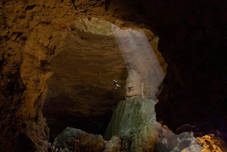 a photograph of an abseiler in a deep cave, dropping down to see a large Buddha statue